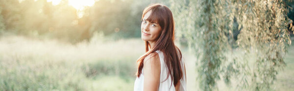 Beautiful happy smiling middle age young Caucasian woman with long dark brown red hair. Casual style real woman female in long white dress outdoor in park nature. Web banner header.
