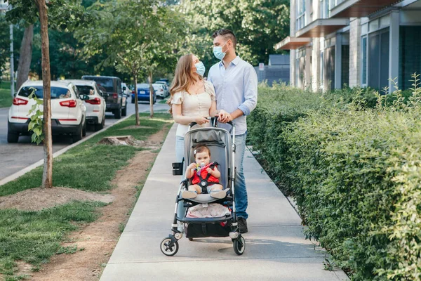 Caucasian mother and father in face masks walking with baby in stroller. Family strolling together outdoors on city street on summer day. New normal in coronavirus covid-19 pandemic.