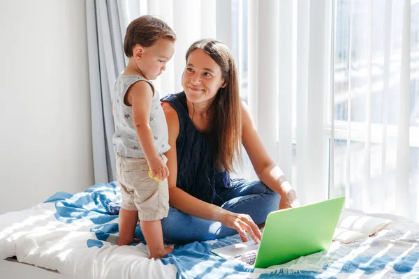 Work from home with kids. Mother working on laptop with baby boy toddler at home. Remote work, distant job. Workplace of freelancer woman with kids. Stay home single mom.