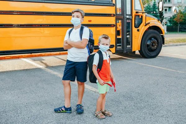 黄色のスクールバスの屋外に立って顔マスクの兄弟学生 顔に個人用保護具付きの子供 教育と9月に学校に戻る コロナウイルスの新常態 — ストック写真