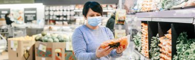 Market alışverişi. Mavi koruyucu maskeli, kısa siyah saçlı orta yaşlı bir kadın marketten sağlıklı sebzeler alıyor. Coronavirus covid-19 'da yeni bir normal. Web başlığı.
