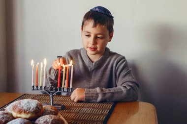 Boy in kippah lighting candles on a menorah for traditional winter Jewish Hanukkah holiday. Child celebrating Chanukah festival of lights at home. Religious Hebrew Judaic Israel culture holiday clipart