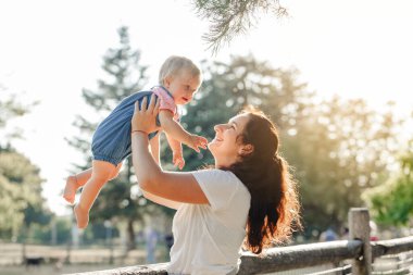Anneler Günü tatili. Genç, gülümseyen beyaz anne ve kız bebek parkta sarılıyorlar. Anne, yaz günü bebeğini dışarıda tutuyor. Mutlu otantik aile çocukluk hayatı. 