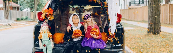 Trick or trunk. Children celebrating Halloween in trunk of car. Boy and girl with red pumpkins celebrating traditional October holiday outdoors. Safe alternative celebration. Web banner header.