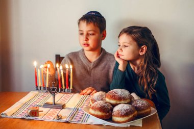 Brother and sister siblings lighting candles on menorah for Jewish Hanukkah holiday at home. Children celebrating Chanukah festival of lights. Dreidel and Sufganiyot donuts in plate on table.  clipart