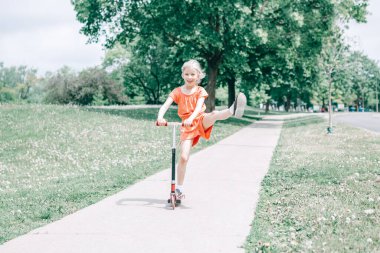 Caddedeki yol parkında kırmızı turuncu tulumlu şirin, komik, beyaz bir kız çocuğu scooter kullanıyor. Çocuklar için yaz eğlencesi çevre sporu aktivitesi. Otantik çocukluk yaşam tarzı.