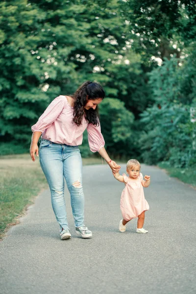 赤ちゃんの第一歩 かわいい赤ちゃんの女の子が歩いて 彼女の母親の手を保持することを学ぶ 幼児の子供の娘が夏の日に屋外の公園に行くのを助けるお母さん 親からのケア サポート — ストック写真