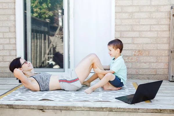 家族スポーツ活動 中年白人の母親で 幼児が裏庭で座り練習をしています 遠く離れた屋外ノートパソコンとオンラインフィットネスワークアウト 子供の少年との親の訓練 — ストック写真