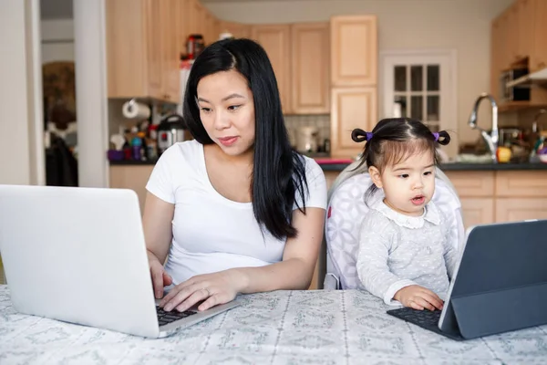 Asian Chinese mother with daughter baby working together on laptop, tablet. Workplace of freelancer woman with kid. Stay home mom working distant job. Online education homeschooling. New normal