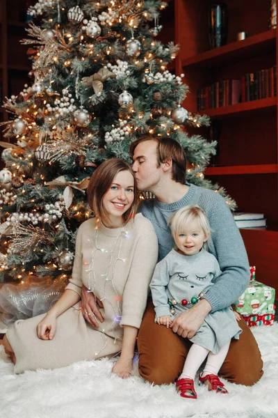 Winter holidays with family. Smiling happy Caucasian mother and father with baby girl daughter by decorated Christmas tree at home. Happy family celebrating Christmas or New Year together.