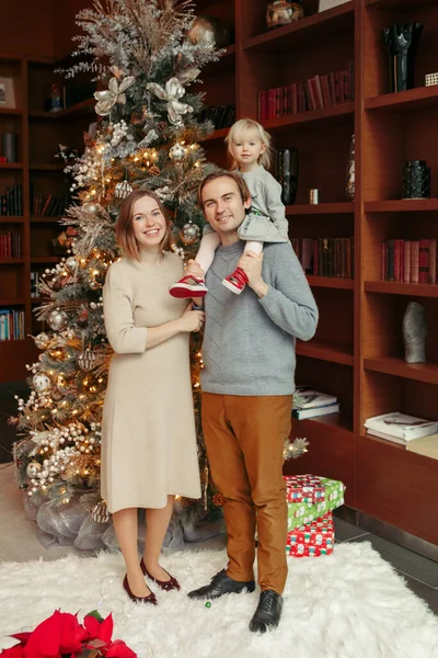 Winter holidays with family. Smiling Caucasian mother and father with baby girl daughter standing by decorated Christmas tree at home. Happy family celebrating Christmas or New Year together.