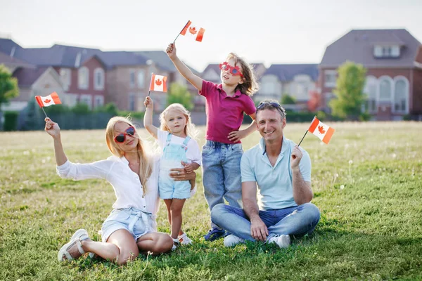 Happy Canada Day. Caucasian family with kids boy and girl sitting on ground grass in park and waving Canadian flags. Parents with kids children celebrating Canada Day on 1st of July outdoor.