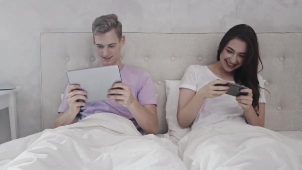 夫妻在床上玩手机游戏。夫妻在桌面上玩和电话。夫妇在床上使用电话和表格游戏。早上时间。被红光射中 — 图库视频影像