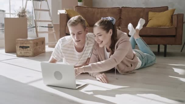 Coppia sdraiata sul pavimento a guardare il computer portatile che parla e indica sullo schermo. Coppia Trasferito nella nuova casa e utilizzando il computer portatile. Shopping online.Hand Motion girato su RED — Video Stock