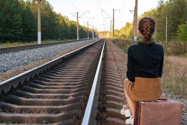 Girl in vintage clothes sits on a vintage shabby brown suitcase on the rails and looks into the distance at the railway, stretching into the forest. Sadness of canceled travels and trips. Copy space