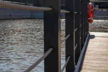 Modern tarz bir ip ve dalgalı mavi suyun yanında ahşap bir çit. Gri tahta döşeme, bağlı can simidiyle birlikte güvenlik çiti. Bir Avrupa şehrinde suya tam donanımlı erişime yakından bak