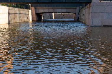 Taşlarla süslenmiş bir su kanalı manzarası. Baharda şehir parkında geniş nehir suyu kanalı. Gölgeli güneş ışığının güzel yansımaları. Çağdaş beton köprüsü olan taştan bir set.