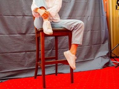 Beyaz gömlekli, mavi kotlu ve beyaz spor ayakkabılı tanınmayan genç bir kız, stüdyo içinde arka planda gri kumaşla yüksek bir bar taburesinde oturuyor. Kollarını bacak üstüne at, yan gözle bak.