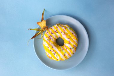 Beyaz, sarı kremalı ve sarı bir tabağa serpiştirilmiş küçük renkli çiçeklerle donut. Mavi arkaplan.