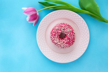 Pembe donut üzerine beyaz çikolata serpilmiş ve pembe bir tabağa yerleştirilmiş. Tabağın etrafında pembe bir lale çiçeği var. Güzel mavi arkaplan.