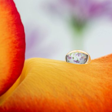 Bir gül yaprağının üzerine küçük bir damla su bırakıldı. Damlada küçük mor bir çiçek resmi var..