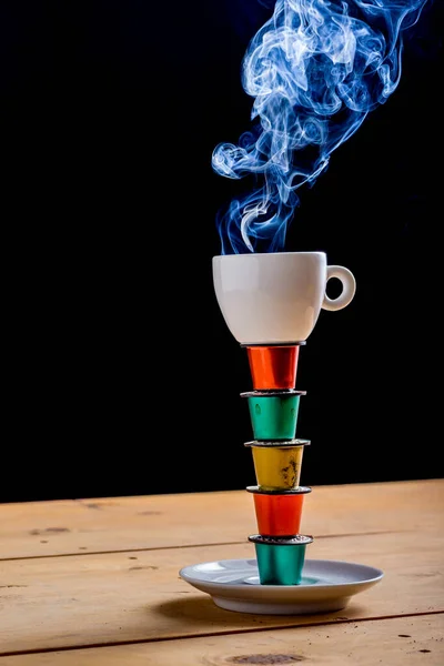 Cup of coffee on top of a tower made with colored coffee pods and used on wooden table on black background.