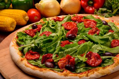 Arugula ve güneşte kurutulmuş domates pizzası tahtada ve sebzeler arka planda..