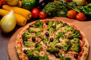 Tahtada siyah zeytinli brokoli pizza ve arka planda sebze..