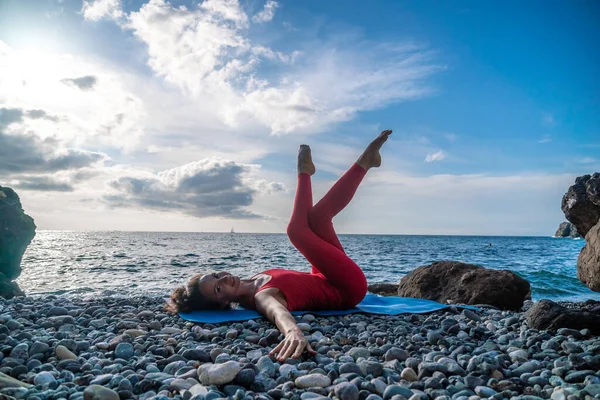 The woman in a red suit practicing yoga on stone at sunrise near the sea. Young beautiful girl in a red bathing suit sits on the seashore in lotus position. Yoga. Healthy lifestyle. Meditation