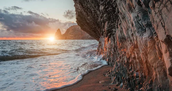 Bright sea sunset. The waves crash into the rock, lit by the warm sunset, sand and pebbles, volcanic basalt as in Iceland. Sea wave breaks into splashes and white foam. Never-ending beauty of nature