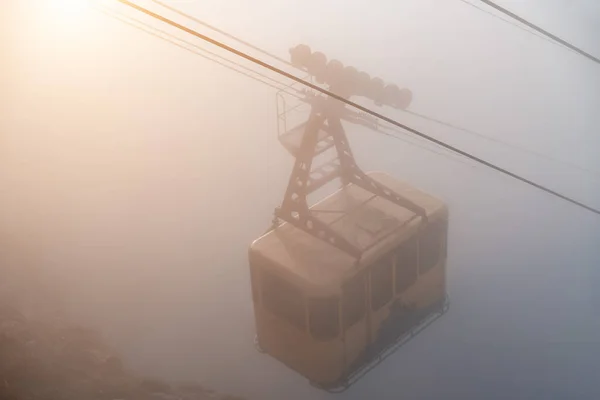 The cable car in Crimea Ai-Petri mountains in thick fog. View from the cliff to the cable car with a rising funicular, mountains in the clouds illuminated by the bright sun. Outdoors travel concept.