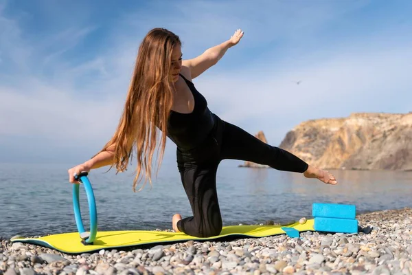 Young woman with long hair, fitness instructor in black Sportswear Leggings and Tops, stretching on a yoga mat with magic pilates ring near the sea on a sunny day, female fitness yoga routine concept