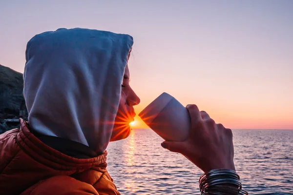 A young tourist Woman holding coffee tumbler cup while sitting outdoor and enjoying sunrise over sea mountain landscape. Womens yoga fitness routine. Healthy lifestyle, harmony and meditation