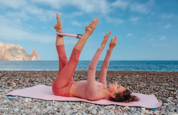 Young woman with black hair, fitness instructor in pink sports leggings and tops, doing pilates on yoga mat with magic pilates ring by the sea on the beach. Female fitness daily yoga concept.