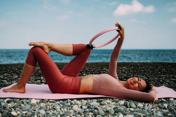 Young woman with black hair, fitness instructor in pink sports leggings and tops, doing pilates on yoga mat with magic pilates ring by the sea on the beach. Female fitness daily yoga concept.