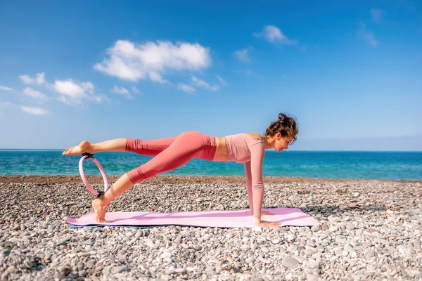 Young woman with black hair, fitness instructor in pink sports leggings and tops, doing pilates on yoga mat with magic pilates ring by the sea on the beach. Female fitness daily yoga concept.