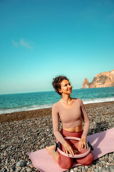 Young woman with black hair, fitness instructor in pink sports leggings and tops, doing pilates on yoga mat with magic pilates ring by the sea on the beach. Female fitness daily yoga concept.