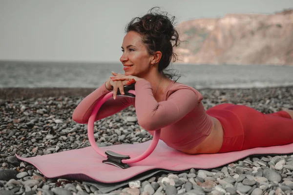 Young woman with black hair, fitness instructor in pink sports leggings and tops, doing pilates on yoga mat with magic pilates ring by the sea on the beach. Female fitness daily yoga concept.