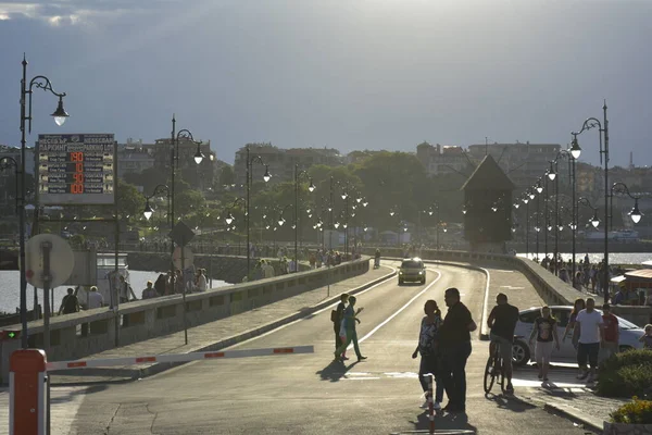 Emberek sétálnak át az utcán naplementében. Egy autó közeledik az autóhídon, az utcai lámpák mintha napnyugtakor ragyognának.. — Stock Fotó
