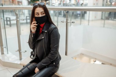 Alışveriş merkezinde siyah maskeli ve cep telefonu olan bir kız. Koronavirüs salgını. Maskeli bir kadın alışveriş merkezinde duruyor. Koruyucu maskeli bir kız alışveriş merkezinde alışveriş yapıyor..