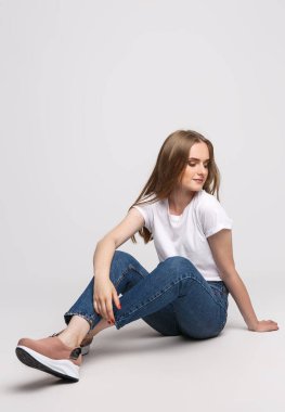Beyaz tişörtlü ve beyaz arka planda kot pantolonlu güzel bir kadın. Genç kadın beyaz arka planda oturuyor. Kıyafet ve ayakkabı reklamları için fotoğraf konsepti.