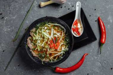 Asya mutfağı, arka planda siyah tabakta sığır eti çorbası. Baharatlı biftek çorbası, sığır filetosu, udon eriştesi, kırmızı biber ezmesi, Pekin lahanası, acı biber, kişniş, seçilmiş sos.