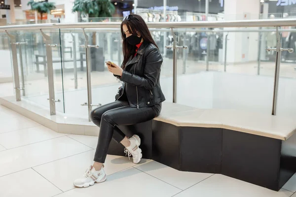 Girl with medical black mask and mobile phone in a shopping center. Coronavirus pandemic. A woman with a mask is standing in a shopping center. A girl in a protective mask is shopping at the mall.