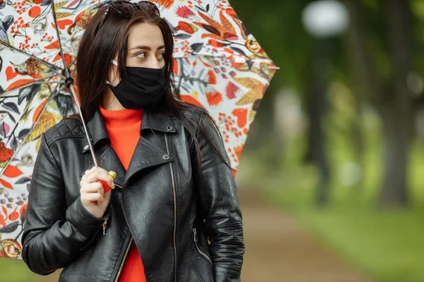 Po ulici kráčí maskovaná dívka. Dívka v ochranné masce chodí do parku s deštníkem v dešti. Koronavirová infekce COVID-19 — Stock fotografie