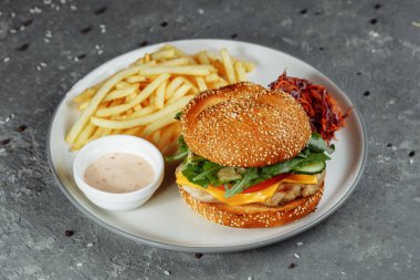 Peynirli hindi burger, marul, domates ve taze salatalık. Gri bir arka planda kızarmış patates ve hindili pirzola.