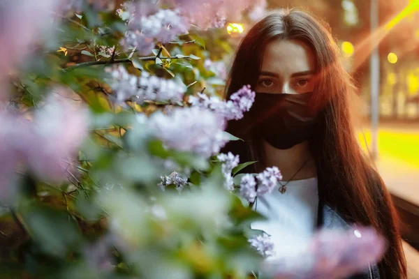 Fille dans un masque médical sur un fond de lilas en fleurs. Masque noir. Protection contre le virus, la grippe. Protection contre le coronavirus. épidémie de coronavirus. l'arôme un arbre dans le jardin Allergie printanière — Photo