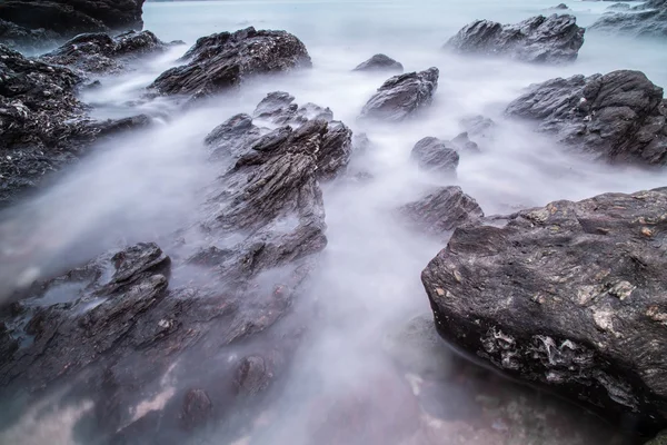 Morbide onde di oceano al tramonto con pietre sullo sfondo della spiaggia. Colpo di lunga esposizione al paesaggio marino — Foto Stock