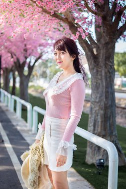 Pembe tişörtlü mutlu genç kız Asya 'da bahar aylarında tatildeyken arka planda kiraz ağaçlarıyla eğleniyor ve dinleniyor. Seyahat kavramı