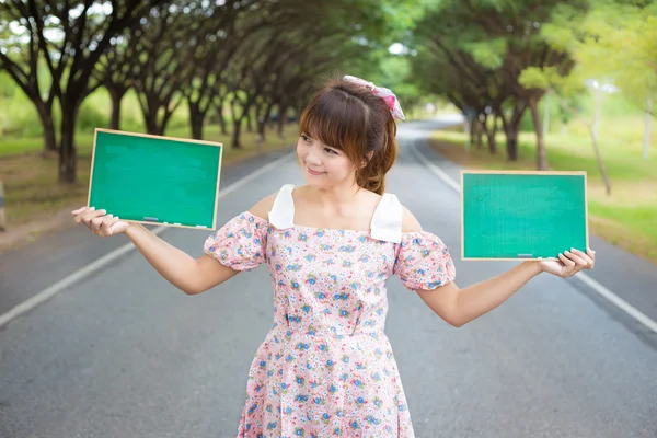 Linda mano de mujer sosteniendo letrero de tablero en blanco verde en el camino con árbol, modelo femenino sonriente . —  Fotos de Stock