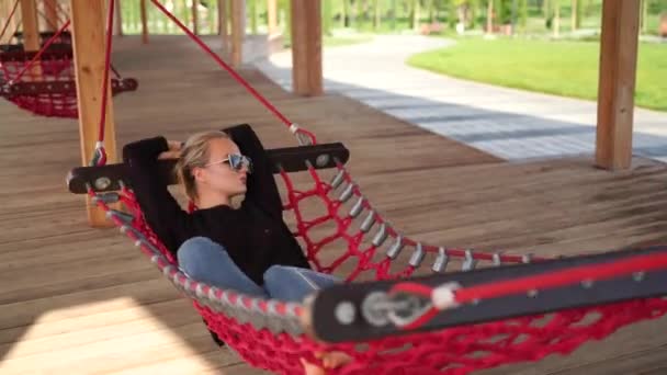 Bella bionda felice si trova su un'amaca e cavalca. Il hammock è situato in un parco della città. Il concetto di ricreazione urbana. — Video Stock